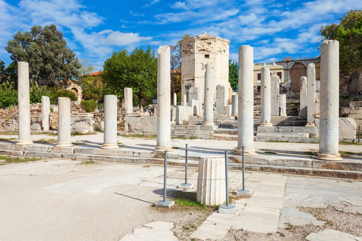 The image shows the Roman Agora in Athens, featuring ancient marble columns and ruins. In the background, the Tower of the Winds, an octagonal structure with carvings, stands among trees and historic buildings. The sky is partly cloudy with blue patches_top 10 places to visit in athens